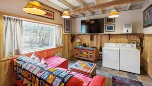 A cozy room with a wooden ceiling, a large window, a colorful blanket on a red sofa, a television, and a washing machine.