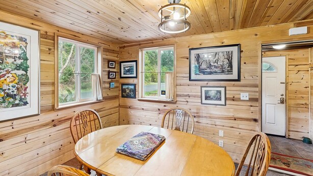 A wooden cabin interior with a round dining table, chairs, framed pictures, and a ceiling light fixture.