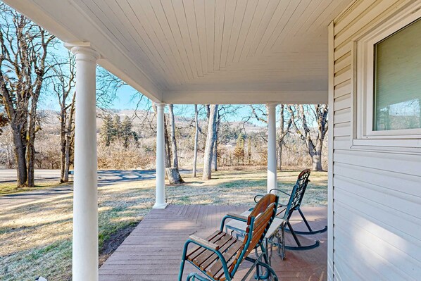A covered porch with two rocking chairs, a wooden bench, and a view of a park with trees and a distant hill.