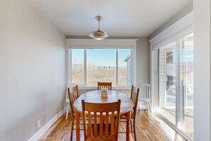 A dining area with a round table, six wooden chairs, a hanging light fixture, and large windows offering a view of the outdoors.