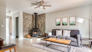 A living room with a stone wall, fireplace, sofa, coffee table, and a ceiling fan. - Spacious Mountain Home with Decks, Firepit, Grill, Wood Stove, W&D, & Free WiFi (Rhododendron)