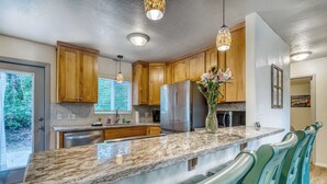 A modern kitchen with wooden cabinets, a stainless steel refrigerator, a marble countertop, and a green bar stool. - Single-level, dog-friendly home w/ private fenced-in yard & easy beach access (Bandon)