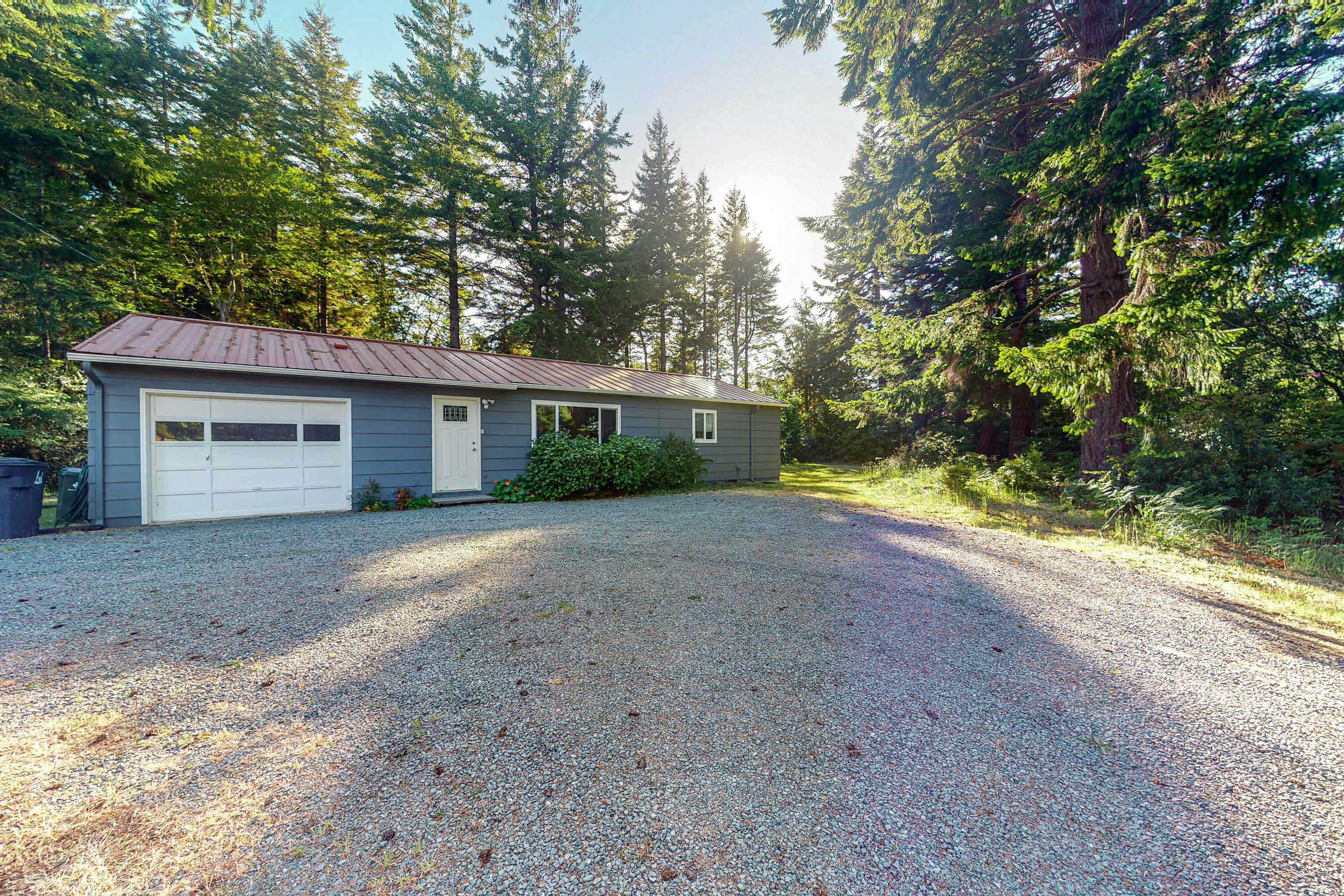 A single-story house with a gravel driveway, surrounded by tall trees.