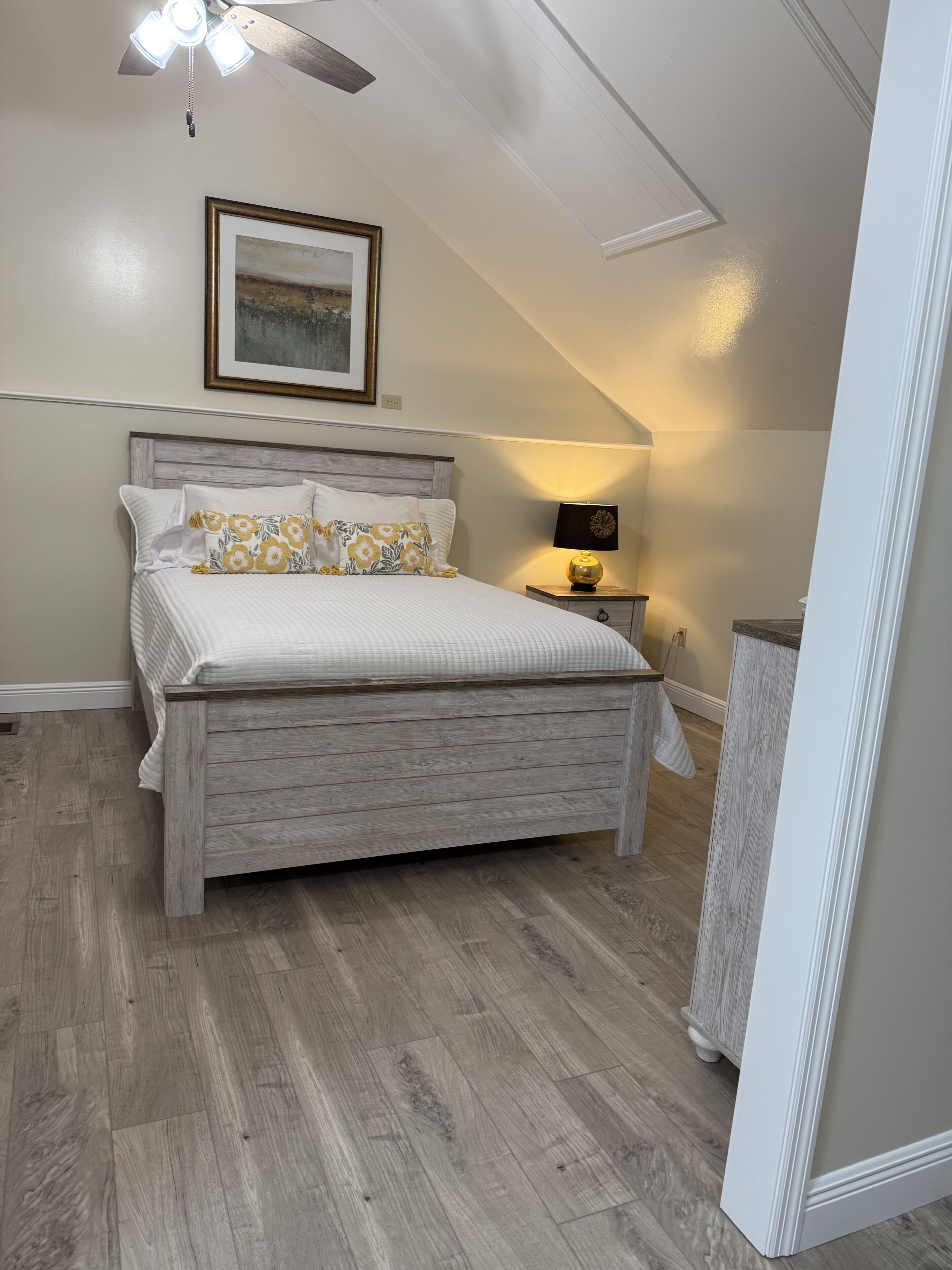 Une chambre à coucher avec un lit en bois, un ventilateur de plafond, un tableau encadré et une lampe de chevet.