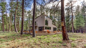 A modern cabin surrounded by tall pine trees, with a deck and outdoor seating area. - Unique & secluded forest home with pool, hot tubs, tennis, sauna, & basketball (Black Butte Ranch)
