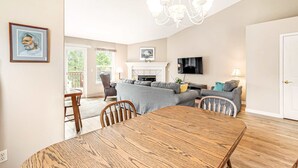 Ein Essbereich mit Holztisch und -stühlen, einem grauen Sofa, einem Fernseher und einer Chandelüster.
