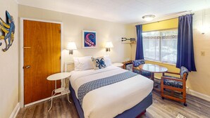A hotel room with a bed, bedside table, chair, lamp, artwork, and a window with curtains. - Retro getaway near beach with fast WiFI - walk to downtown shops & eateries (Bandon)