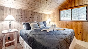 Une chambre à coucher aux murs lambrés, avec un lit, deux lampes de chevet et une fenêtre équipée de stores.