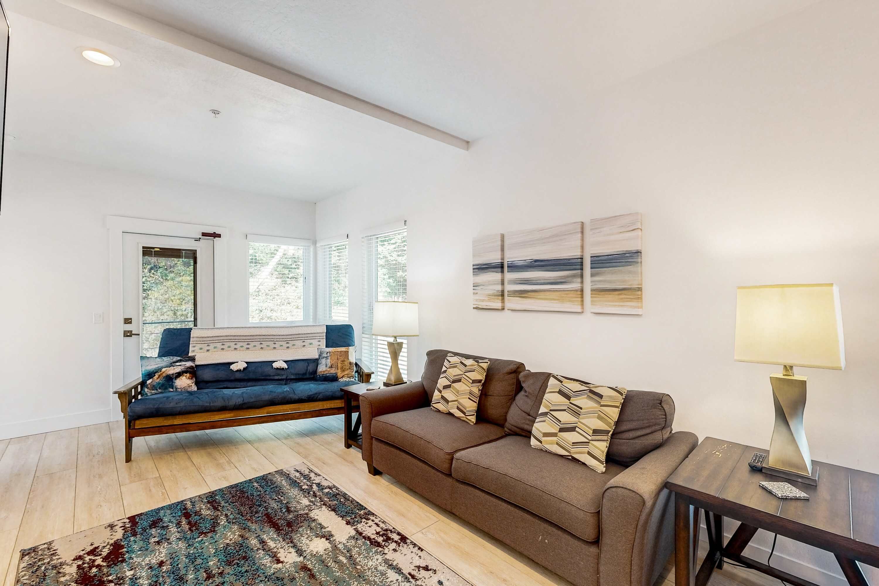A living room with a sofa, a coffee table, and a lamp. There are windows and artwork on the walls.