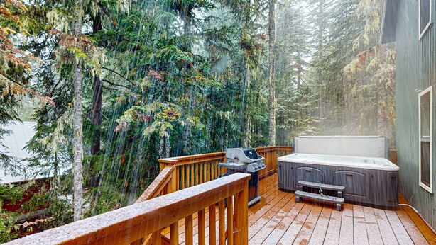 Eine Holzterrasse mit Whirlpool und Grill, umgeben von Bäumen und Blick auf einen See.