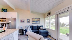 A modern kitchen with a refrigerator, microwave, and a wooden countertop. There is a blue sofa with patterned cushions and a beige armchair. The room has large windows with a view of a green lawn and a building in the distance. - Oceanfront getaway close to Gearhart Golf Links , no pets (Gearhart)