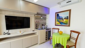 A modern living room with a flat-screen TV, a dining table with chairs, a kitchen area with a built-in oven, and a painting on the wall. - Studio Beira-mar com Vista no Jardim Atl Ntico (Ilhéus)