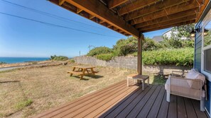 A wooden deck with a picnic table and a small table, overlooking a grassy yard with a view of the ocean. - Newly Remodeled Ocean View Retreat, Balcony and BBQ (Port Orford)
