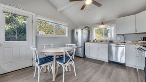 A modern kitchen with white cabinetry, a stainless steel refrigerator, and a dining area with a round table and white chairs. - Newly Remodeled Ocean View Retreat, Balcony and BBQ (Port Orford)