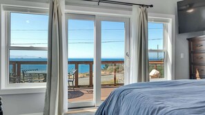 A bedroom with a large bed, a view of the ocean through floor-to-ceiling windows, and a wooden deck outside. - Newly Remodeled Ocean View Retreat, Balcony and BBQ (Port Orford)