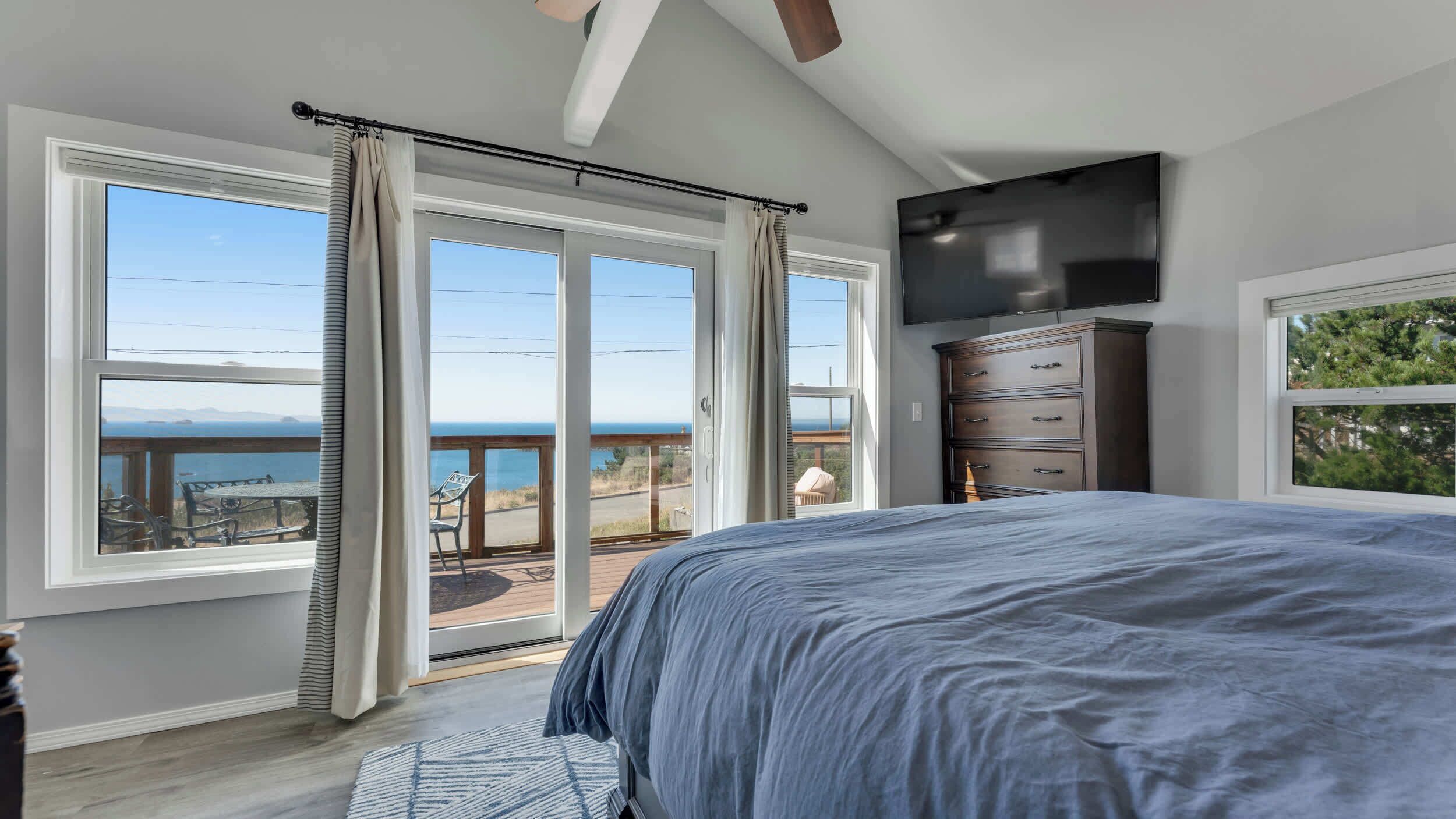 A bedroom with a large bed, a dresser, and a TV. There are three windows with curtains, and a balcony with a view of the ocean.