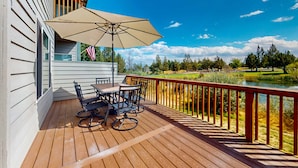 A wooden deck with a table and chairs, a large umbrella, and a view of a golf course. - Charming waterfront townhouse with fireplace, pool, hot tub, & washerdryer (Eagle Crest)