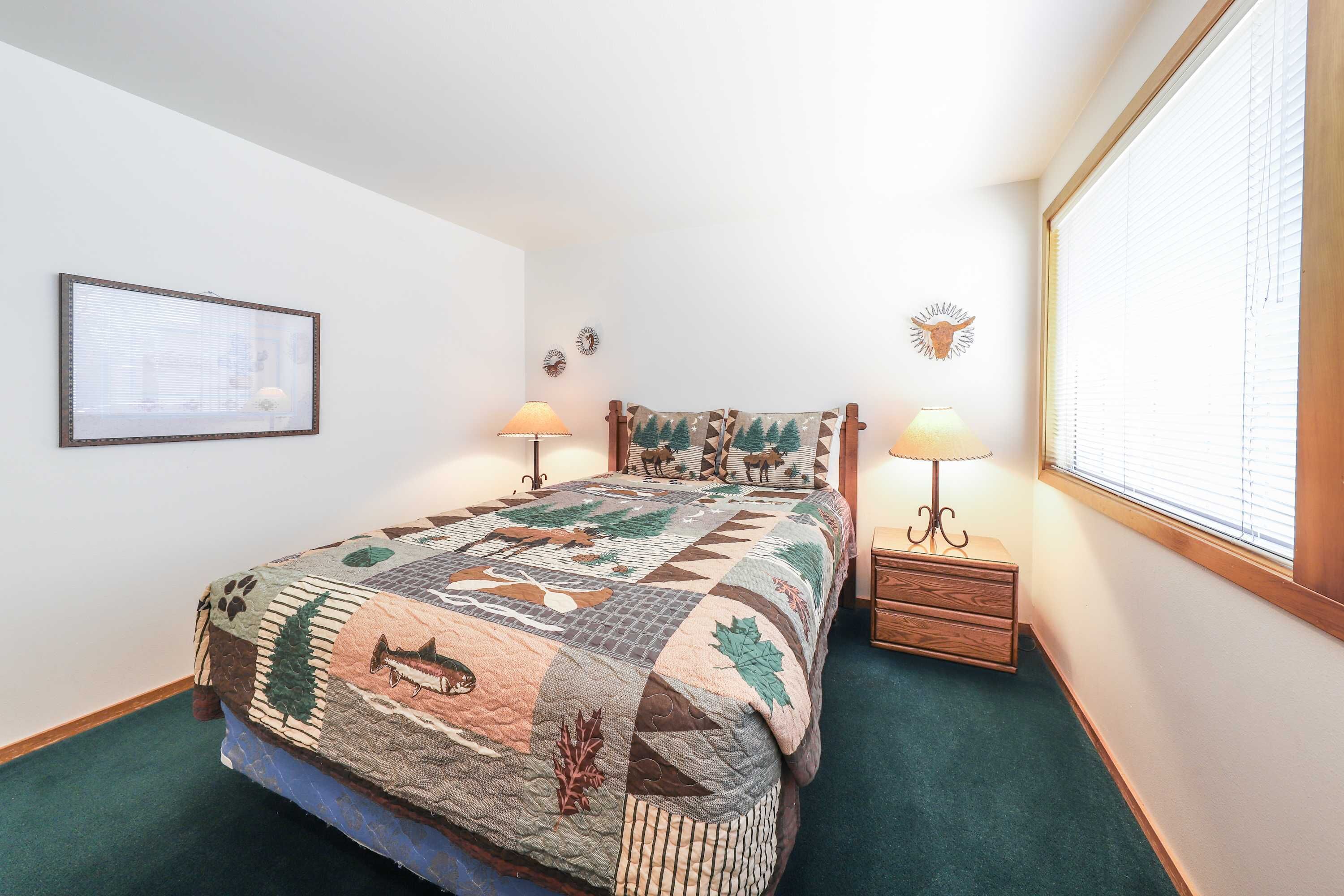 A bedroom with a bed, two bedside lamps, a window with blinds, a framed picture on the wall, and a small wooden nightstand.