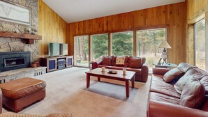 Een ruime woonkamer met een stenen openhaard, houten wanden, een leren bank, een bijzettafeltje en grote ramen met uitzicht op bomen.