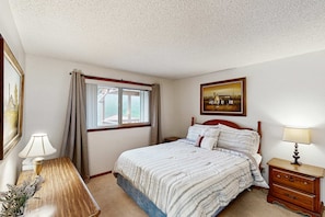 A bedroom with a bed, a nightstand, a lamp, a window with curtains, a painting on the wall, and a vase with flowers.