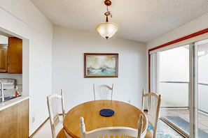 A dining area with a round table, four chairs, a ceiling light fixture, a framed painting, and a view through a glass door.