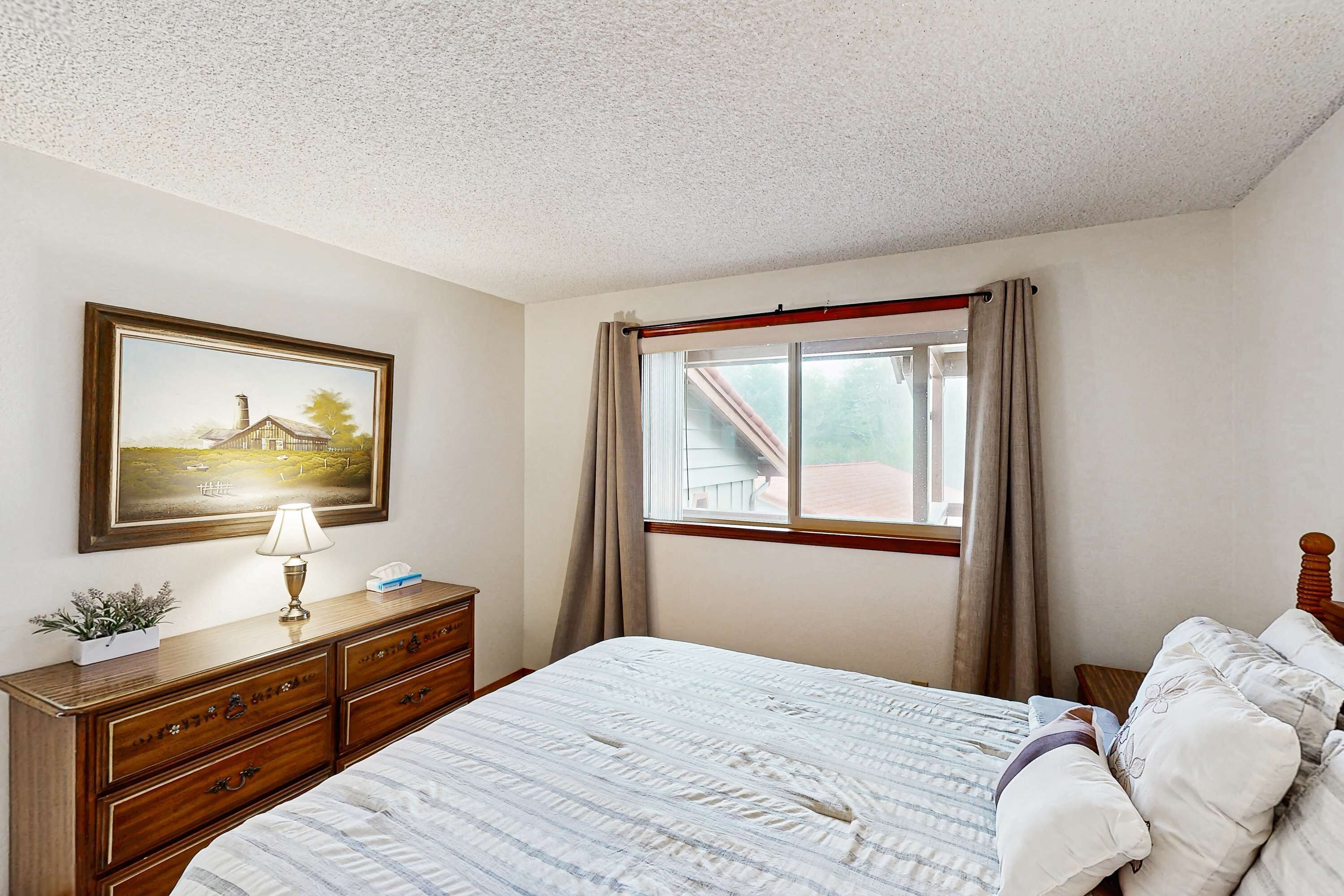 A bedroom with a bed, a dresser, a window with curtains, a painting on the wall, and a lamp.