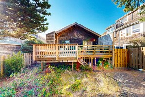En træhytte med terrasse, omgivet af en indhegnet have med beplantning.
