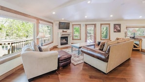 A spacious living room with a fireplace, large windows, and a balcony. - Lovely home with pool, hot tub, balcony, & washer/dryer (Sunriver)