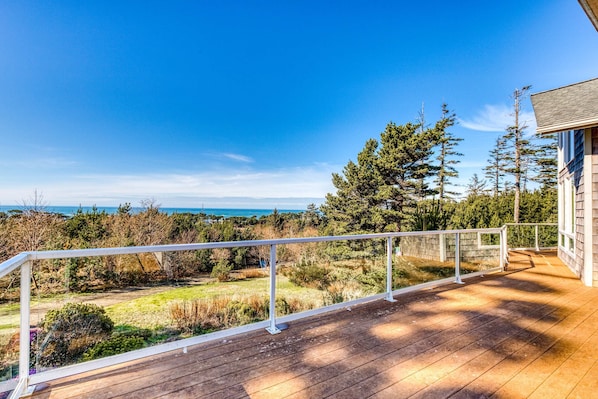 Une terrasse en bois dotée d’une rambarde, qui offre une vue sur une zone verdoyante avec des arbres et, au loin, sur l’océan.
