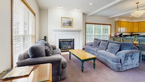 A living room with a fireplace, two grey sofas, a wooden coffee table, and a dining area with wooden cabinets and a chandelier. - Large home 1 block from the beach with ocean view, game room, fireplace & WiFi (Seaside)