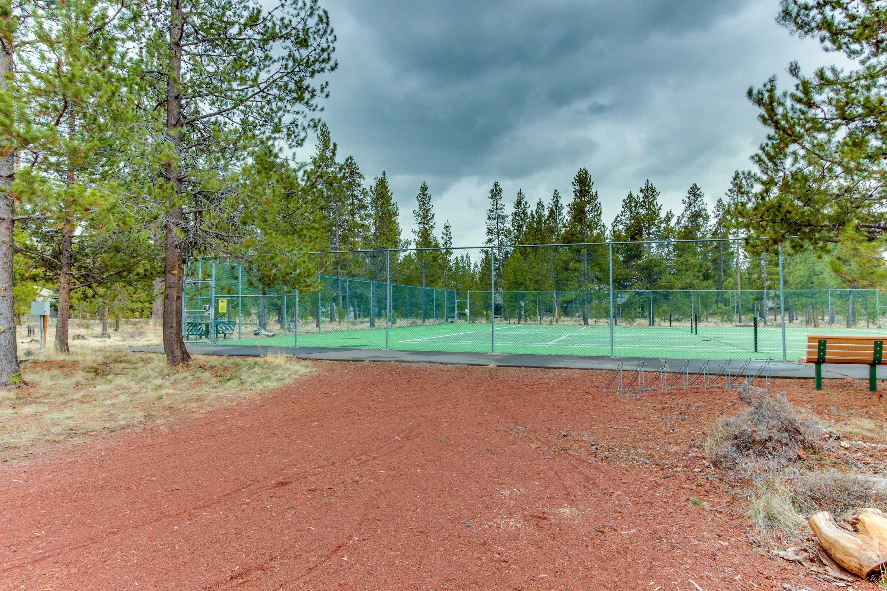Una cancha de tenis con valla verde, un banco y árboles al fondo.