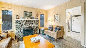 A cozy living room with a stone fireplace, a sofa, a coffee table, and a kitchen visible through an open door. - Charming dog-friendly cottage with sauna, easy beach access, deck & yard (Gearhart)