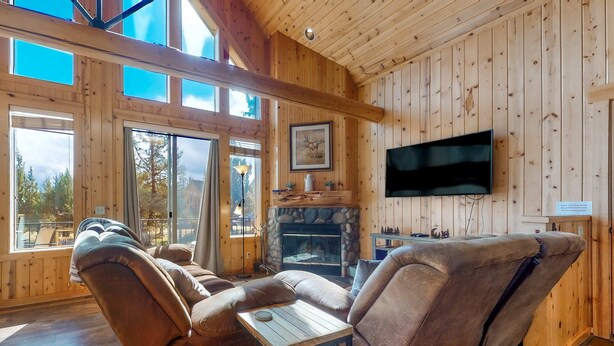 Interior de cabana de tronco com lareira, televisão na parede e uma grande janela com vista para uma floresta.