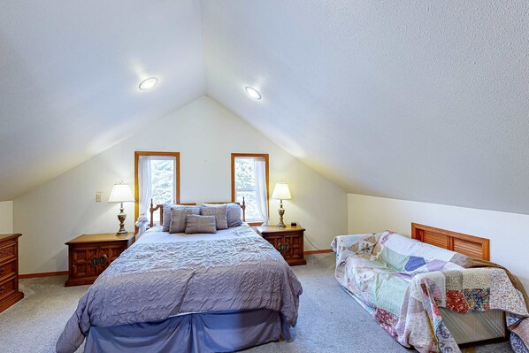 Une chambre avec un plafond en pente, un lit recouvert d’une courtepointe, deux tables de chevet avec des lampes et une fenêtre avec des rideaux.