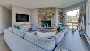 A modern living room with a stone fireplace, a large grey sofa, a flat-screen TV, and a view of the outdoors. - Mountain-view condo on the golf course with WiFi, private W/D & gas grill (Welches)