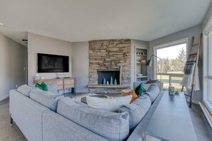 A modern living room with a stone fireplace, a large grey sofa, a flat-screen TV, and a view of the outdoors.