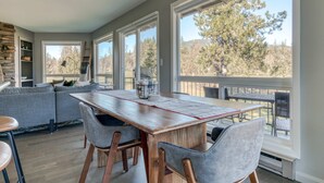 A dining area with a wooden table and chairs, a sofa, and large windows with a view of trees. - Mountain-view condo on the golf course with WiFi, private W/D & gas grill (Welches)