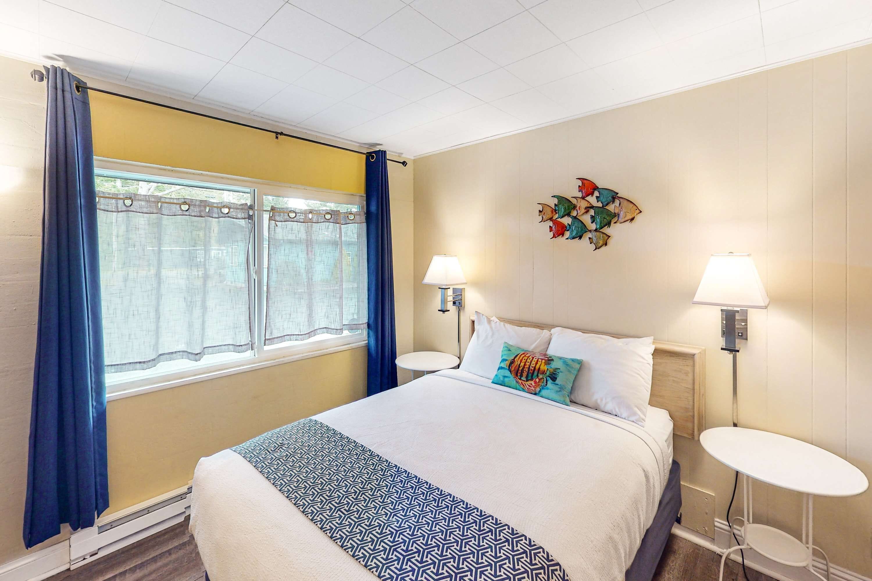Habitación de hotel con cama, dos mesitas de noche, una ventana con cortinas, una obra de arte colgada en la pared y una lámpara de pared.