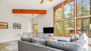 A modern living room with a large gray sectional sofa, a flat-screen TV, and large windows with a view of trees. - House in Black Butte Ranch with shared amenities (Black Butte Ranch)