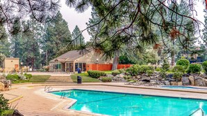 A swimming pool with a surrounding deck, a small building with a red roof, and a backdrop of tall trees. - Mountain view condo with a fireplace, WiFi, partial AC, shared hot tub & pool (Bend)