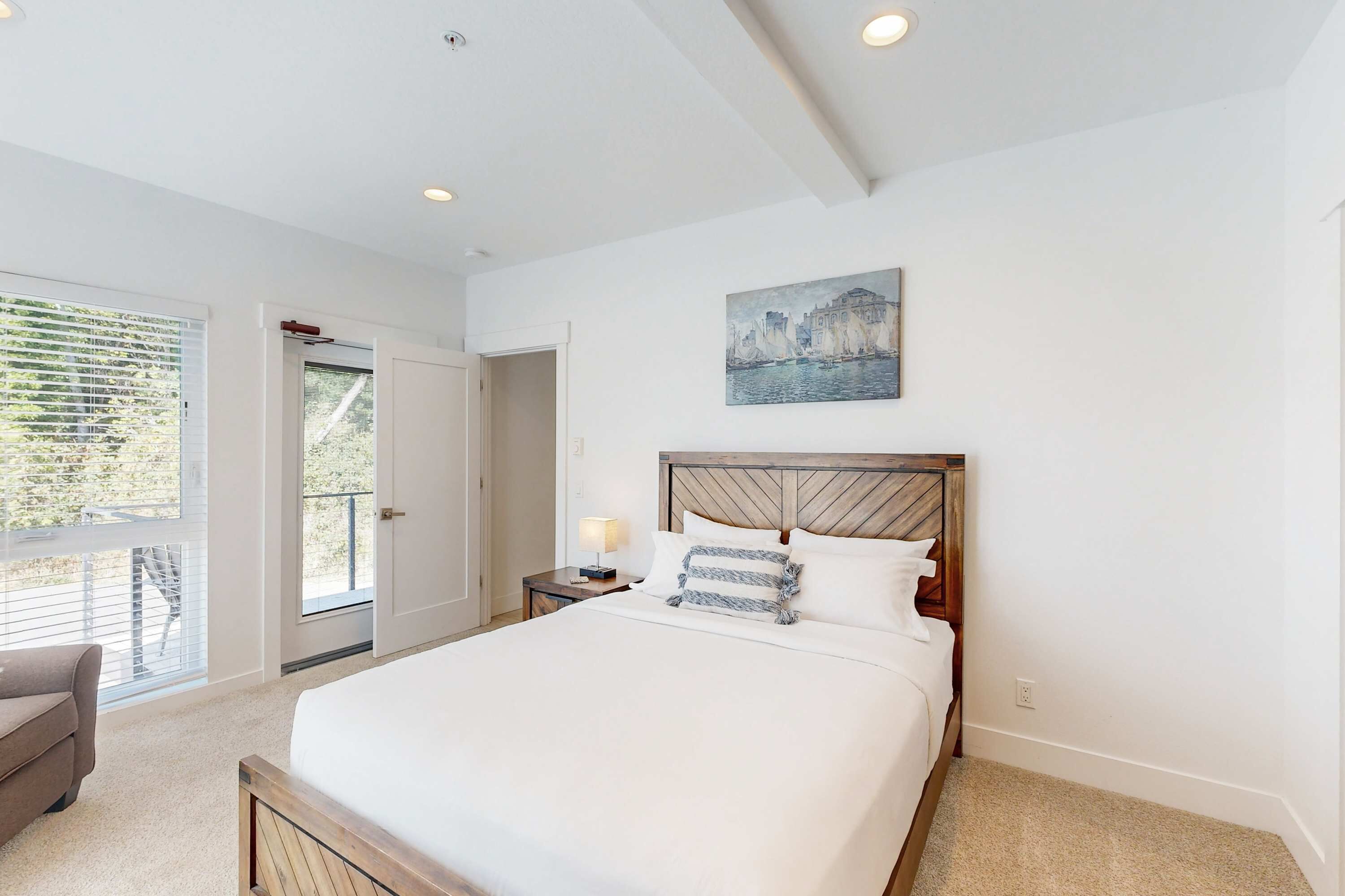 A bedroom with a large bed, a nightstand, a lamp, a painting on the wall, and a door leading to an outdoor area.