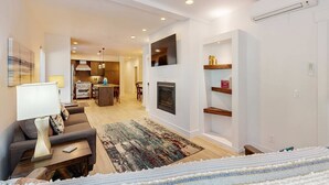 A modern living room with a sofa, coffee table, fireplace, and a kitchen in the background. - Stunner apt with fireplace, grill, partial ocean views & partial AC (Gold Beach)