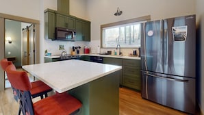 A modern kitchen with green cabinets, a stainless steel refrigerator, and a central island with red chairs. - Amazing Riverfront Cabin with Views, Deck, Firepit, & Grill - Dog-Friendly (Vida)
