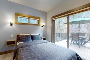 A bedroom with a bed, bedside tables, a window with blinds, and a balcony with outdoor furniture. - Amazing riverfront cabin with views, deck, firepit, & grill - dog-friendly (Vida)