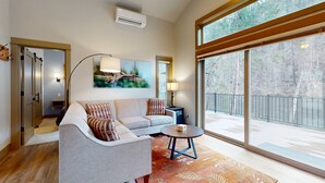 A modern living room with a sofa, a coffee table, a painting, and a large window with a view of trees. - Amazing Riverfront Cabin with Views, Deck, Firepit, & Grill - Dog-Friendly (Vida)