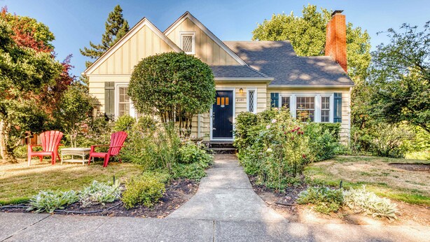 A two-story house with a front porch, a pathway leading to the door, and a small garden with plants and a tree.