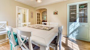 A dining area with a white table, chairs, and a chandelier. There is a kitchen visible through an archway, and a cabinet with glass doors.