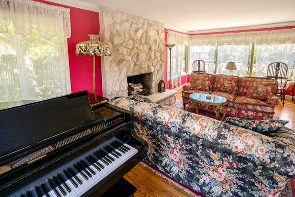 A living room with a grand piano, a stone fireplace, floral-patterned sofas, and a round coffee table. - Classic & elegant house with entertainment & spacious, laid-back yard (College Place)