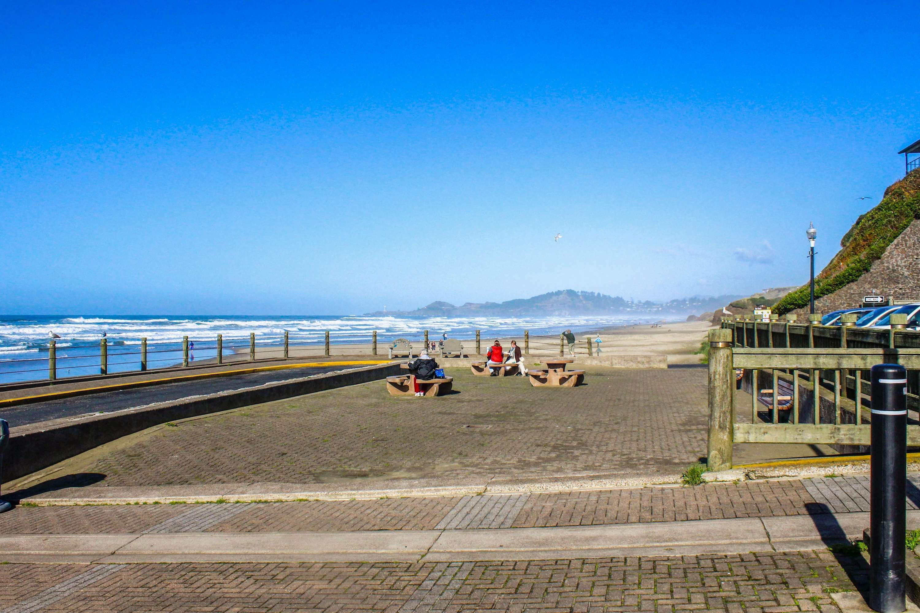 A coastal walkway with picnic tables, a stone wall, and a view of the ocean.