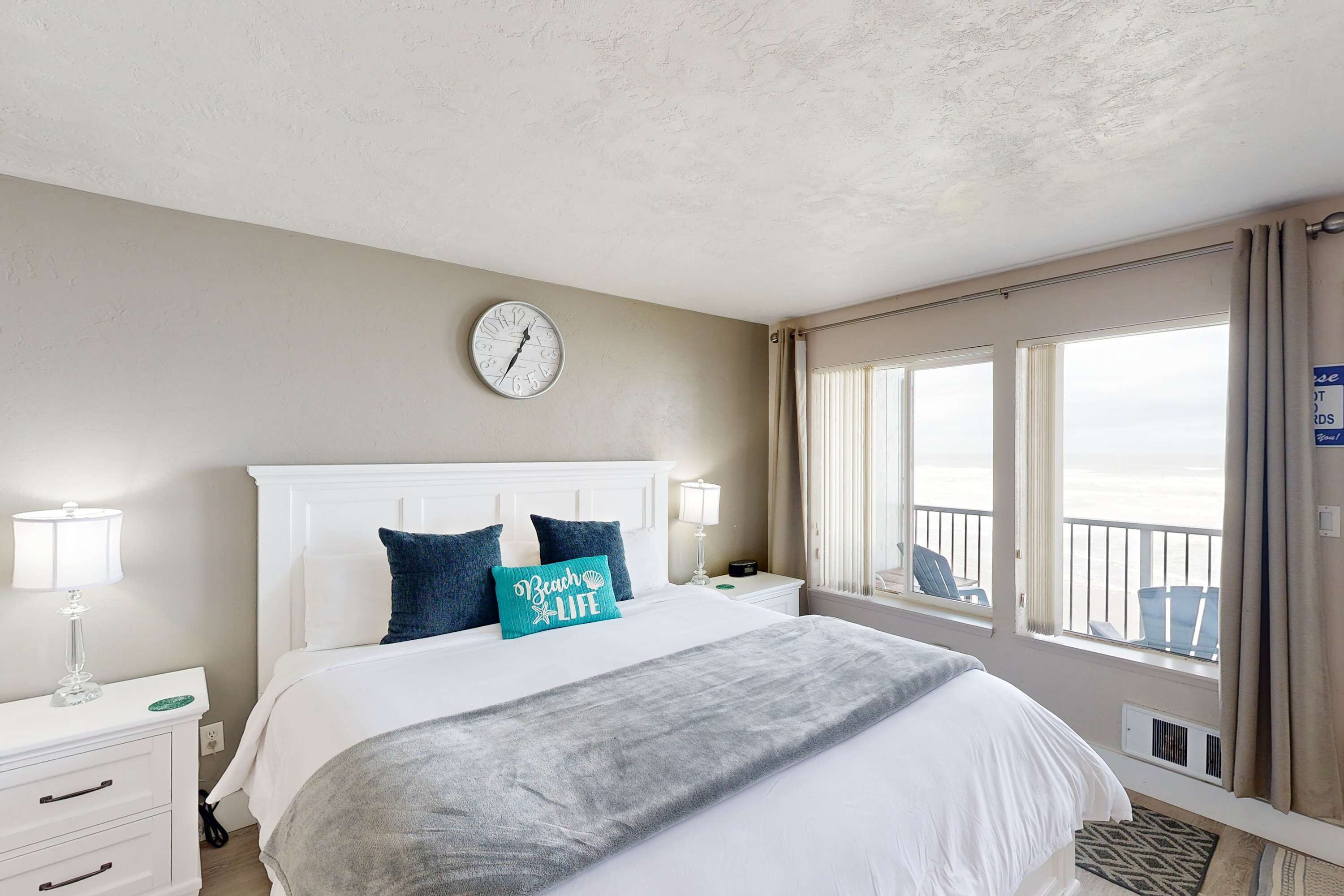A hotel room with a bed, a clock, a window with a view, and a nightstand with a lamp.